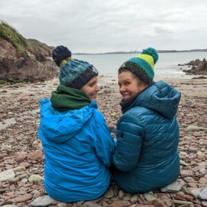 2 smiling friends wearing woolly hats sat on a pebbly beach enjoying a Wild swim & yoga retreat in Pembrokeshire